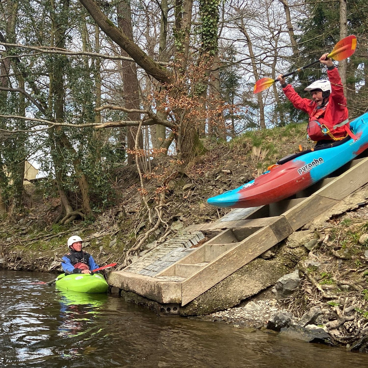 Two people in kayaks at the edge of a wooden platform with trees in the background